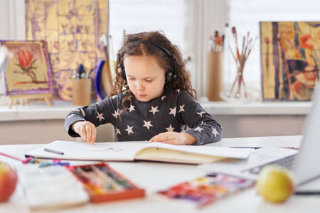 Cute child girl sitting at a table in earphones drawing using watercolor. Homeschooling or art...