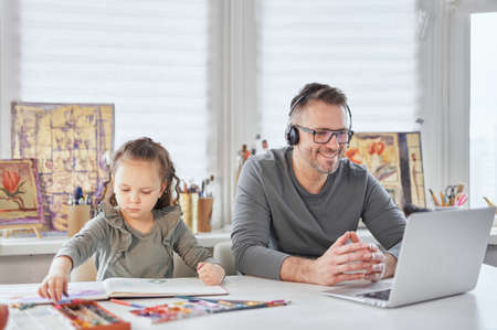 Young caucasian family busy working in their room at the table. Child girl drawing next to dad who is working online with laptop and headphones. Babysitting during covid-19 lockdown. Front view.の写真素材