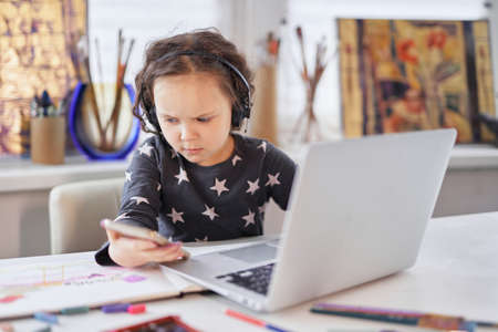 A small kid girl in handsfree headphones sitting at a table using a laptop computer and smartphone....