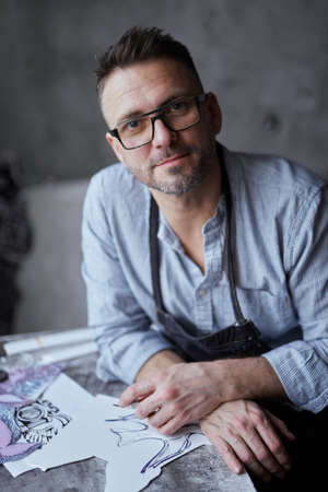 Bearded middle aged male artist in glasses sitting on a background of workplace. Linocut master in...