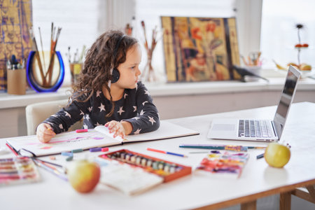 Cute small kid girl in handsfree headphones sitting at a table using a laptop computer for education...