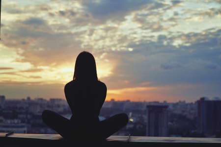 Meditation yoga silhouette of woman at sunset with urban background. Woman practicing yoga during surrealistic sunset with clouds and city view. Mental and physical health, harmony and workout conceptの写真素材