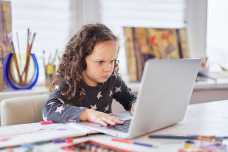 Homeschooling or online studying concept. Cute small child girl sitting at the table in earphones doing homework or online schooling using laptop. High quality photoの写真素材