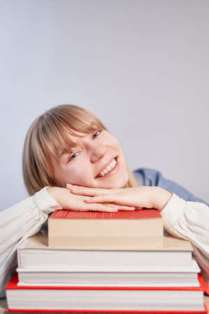 Caucasian blonde student girl relaxing lying on stack of books and smiling. Young female pupil with books in class with copy space. Back to school or homeschooling concept. High quality vertical photoの写真素材