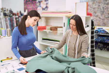 Woman assistant in fabrics shop demonstrating khaki color textile to young tailor girl customer....