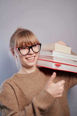 Education or knowledge concept. Thumbs up gesture. Happy positive nerd, attractive university or college blonde student girl wearing eyeglasses and beige sweater holding stack of books in handsの写真素材