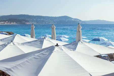 Sunny summer day and white umbrellas in Nice, France. Holiday atmosphere on summer time at French riviera. White umbrellas on a beach in blue clear sky and mountains on background. High quality photoの写真素材