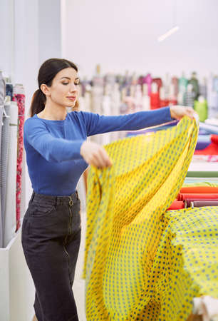 Attractive brunette female customer designer throwing up yellow textile cloth at showroom or store interior. Tailor, seamstress, couture or dressmaker concept. High quality vertical imageの写真素材