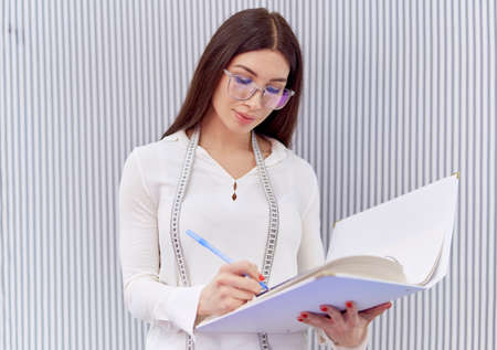 Female stylish brunette warehouse or textile store owner in eyeglasses and measure tape making inventory in a shop. Manager calculating and making notes in journal. Concept of e-commerce or retailの写真素材