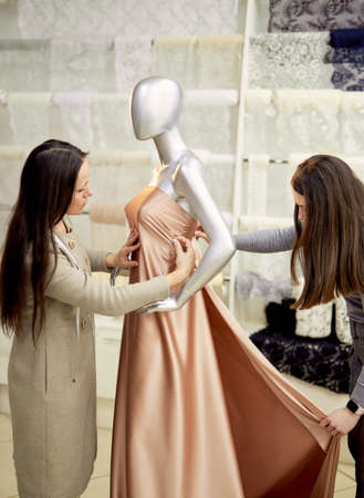 Fashion and tailoring concept. Professional brunette team of dressmakers, designers or seamstresses working with new model applying textile cloth on mannequin or dummy in atelier. High quality imageの写真素材