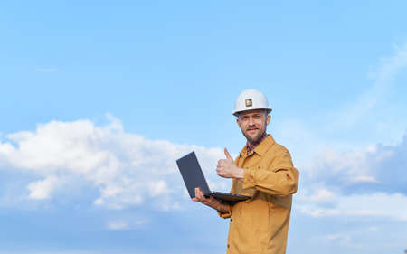 Attractive bearded engineer or construction architect in helmet and orange jacket holding computer notebook in front of sunny day blue sky. Looking at camera and making thumbs up gestureの写真素材