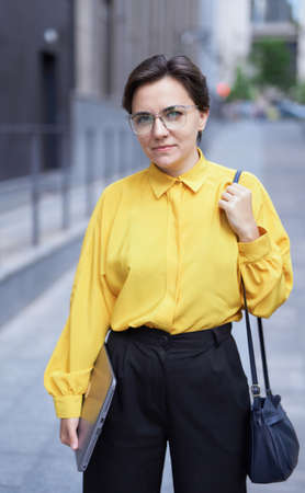 Closeup portrait of attractive businesswoman, manager or finance economist with laptop standing near the office building in downtown wearing yellow blouse and trousers looking straight in cameraの写真素材