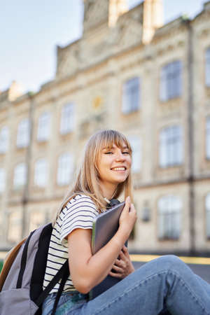 Attractive cheerful young blonde female university or college student with backpack and laptop wearing jeans sitting on green grass in campus. Happy school girl, student portrait or education conceptの写真素材