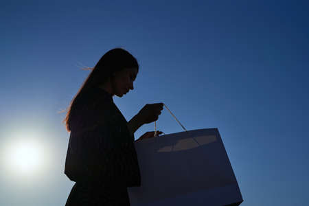 Happy long hair woman looking at shopping bags in sunset, silhouette image against sun and blue sky. Consumerism, shopping, sale or black friday concept. High quality photoの写真素材