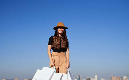 Attractive luxury caucasian woman with shopping bags and blue sky background with city view. Brunette glam girl with shopping bags standing in bright clothes and brown hat. High quality photoの写真素材