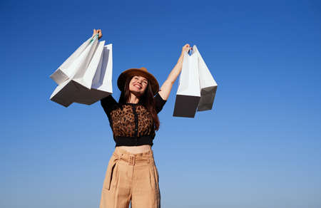 Retail, consumerism or sale concept - happy woman with shopping bags. Attractive shopaholic brunette girl rejoices in shopping on blue sky background holding shopping bags in hand. High quality photoの写真素材