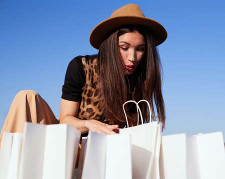 Black friday, shopping, sale or consumerism concept. Glamour beautiful brunette woman in brown hat looking in shopping bags surprised, open mouth amazed expression. High quality vertical photoの写真素材