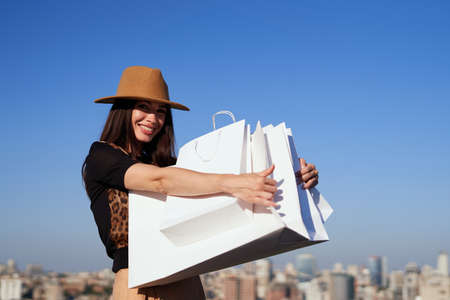 Luxury girl in brown hat holding bunch of shopping bags. Attractive shopaholic brunette girl rejoices in shopping on blue sky background. Retail, black friday or sale concept. High quality photoの写真素材