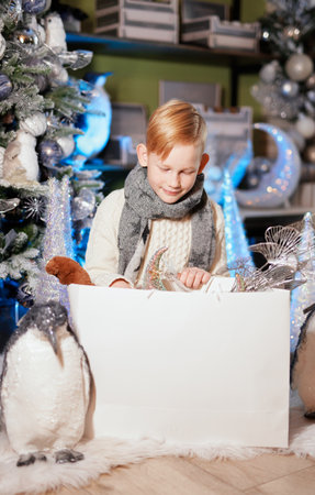 Happy smiling redhead boy wearing scarf and sweater sitting near decorated Christmas tree unpacking New Year decoration in shopping white bag at home with blue light on background. High quality photoの写真素材