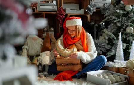 Happy cheerful teenager in Santa hat and red scarf with New Year decoration in wooden box and low...