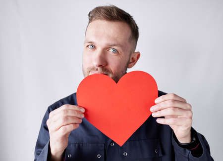 Young bearded male worker standing by the wall holding red heart shape and looking at camera. Worker...