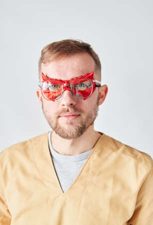 Close up portrait of a male doctor or nurse. Portrait of a handsome caucasian male doctor wearing red face superhero mask, eyeglasses and beige medical uniform. High quality vertical photoの写真素材