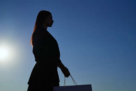 Silhouette of caucasian fashionable long hair woman looking at shopping bag an sunset, image against sun and blue sky. Consumerism, shopping, sale or black friday concept. High quality photoの写真素材