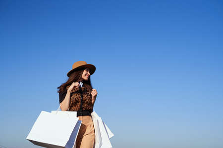 Sale or retail concept. Beautiful caucasian luxury fashionable woman in hat with shopping bags, blue sky and urban city background. Glam brunette girl in sunglasses going outdoor in sunny dayの写真素材