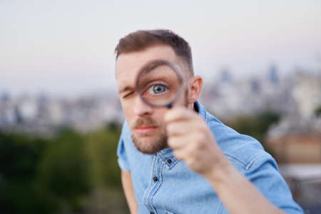 Cute man closeup looking with magnifying glass at camera wearing jeans shirt and peaked cap. Young bearded male as detective, investigator or shopping sales searcher concept. High quality imageの写真素材