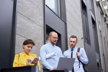 Men and woman in casual clothes standing outside, using laptop, talking and having brainstorming or...