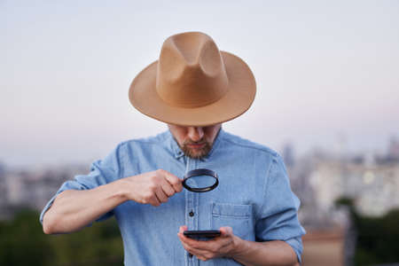 Bearded man looking with magnifying glass at mobile phone. Male wearing jeans shirt and hat looking with loupe at smartphone as detective, investigator or searching concept. High quality imageの写真素材