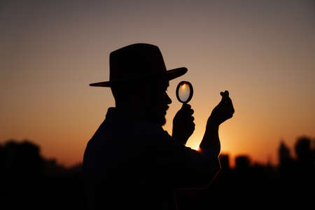 Attractive male silhouette in hat looking with magnifying glass showing small korean heart gesture. Man searching sales or discounts at sunset using loupe with urban view. Investigation conceptの写真素材