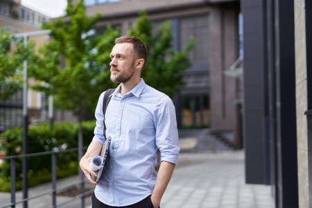 Cute bearded caucasian businessman, real estate agent, designer or engineer standing outdoor with laptop and plans. Modern building and trees on background. High quality imageの写真素材