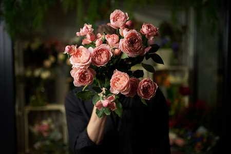 Cute male holding a bunch of fresh pink long stemmed roses in front of his face hiding it. Low key image. Man in black shirt with flower bouquet. Love or romance concept. High quality photoの写真素材