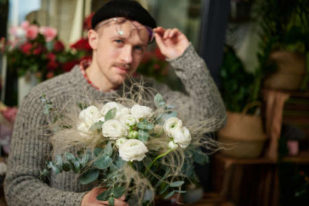 Smiling bearded man with bouquet of white roses in flower shop. Good looking male florist wearing sweater, beanie and eyeglasses holding flower bouquet. Small business concept. High quality imageの写真素材
