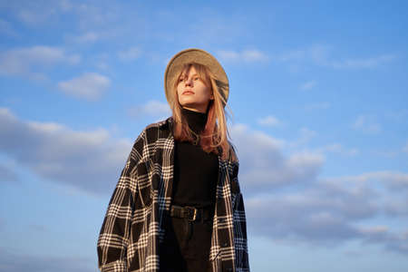 Portrait of a girl looking forward. Attractive blonde long hair woman in stylish clothes - shirt and brown hat on with blue cloudy sky background. High quality imageの写真素材