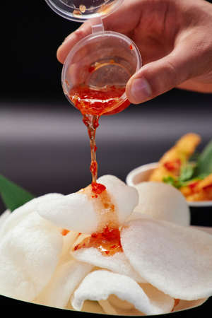 Adding sweet and spicy chili sauce on Chinese chips on a paper plate. Junk food or fastfood concept. Close-up of krupuk chips on black background. High quality photoの写真素材