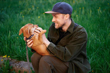 Dog as family member concept. Attractive caucasian man in baseball cap sitting on a grass in park and playing with small brown puppy biting him on finger. Young male with dachshund outdoorsの写真素材