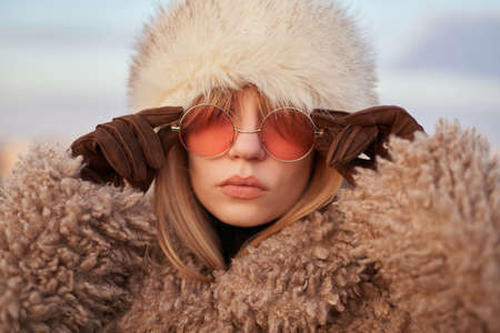 Attractive fashionable glamour young girl in stylish clothes - artificial fur coat and hat correcting pink sunglasses with hands in brown gloves on background of blue cloudy sky. High quality imageの写真素材