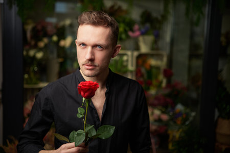 Romantic, bachelor or love concept. Serious sexy masculine macho male with mustache holding one red rose in hand, looking at camera wearing black shirt with dark background.の写真素材