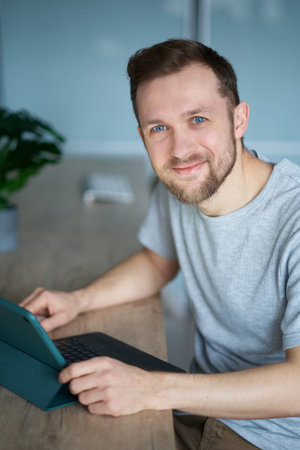 Attractive bearded caucasian male freelance software engineer, developer or web designer sitting at working place in office hardly working using computer looking at camera smiling. High quality imageの写真素材