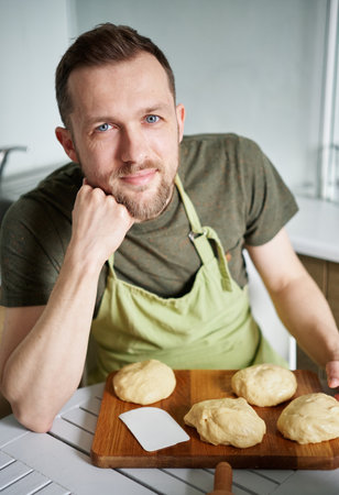 Portrait of caucasian baker man in green apron uniform smiling looking at camera. Portrait of handsome baker at home sitting at the table with dough. High quality vertical photoの写真素材