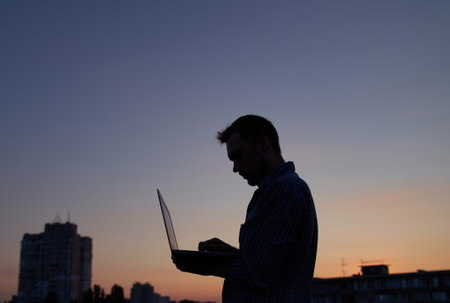 Freelancer developer or man it engineer working outdoor using laptop in silhouette at sunset with buildings. Adorable dusk at background. Hardly working evening. High quality imageの写真素材