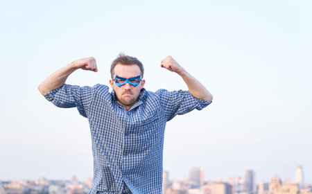 Adorable caucasian man in nightwear shirt and superhero blue mask showing strength gesture and angry...