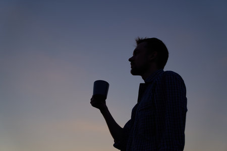 Male person in silhouette outdoor drinking coffee or tea at sunset. Adorable dusk at background. High quality imageの写真素材