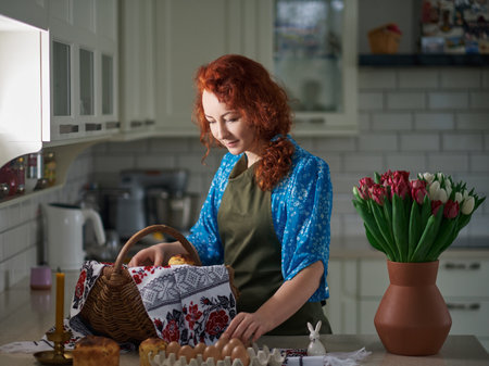Woman smiling, preparing Easter basket with embroidered towel at kitchen home with tulips standing...