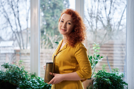 Caucasian redhead florist woman portrait at home with watering can and green plants on background. Florist, plants care and gardening concept. Female in yellow dress. High quality imageの写真素材