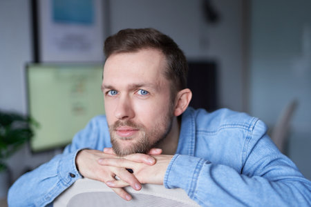 Young handsome man in denim shirt, positive emotion with computer monitor on backgroundSerious bearded male software engineer sitting at working place in office looking at camera. High quality imageの写真素材