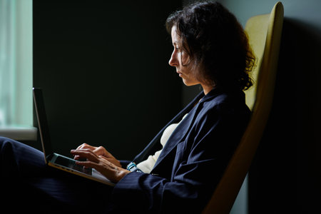 Profile shot of female in business suit looking at laptop during working process in co-working open office space on the dark background. High quality photoの写真素材