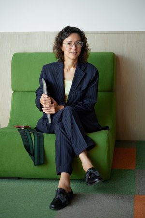 business woman in glasses smiling looking at the camera with laptop sitting in the chair in the office. Dark jacket, confident woman looking in the camera High quality photoの写真素材